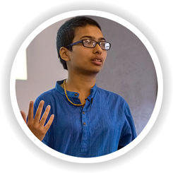 Person gesturing while speaking, wearing a blue shirt and glasses, in a round frame with a blurred background.