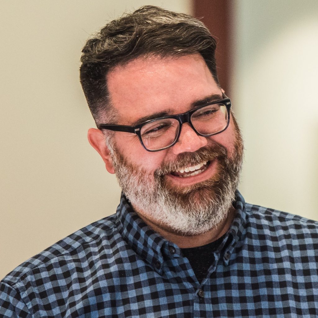A man with glasses and a beard smiling, wearing a blue checkered shirt.