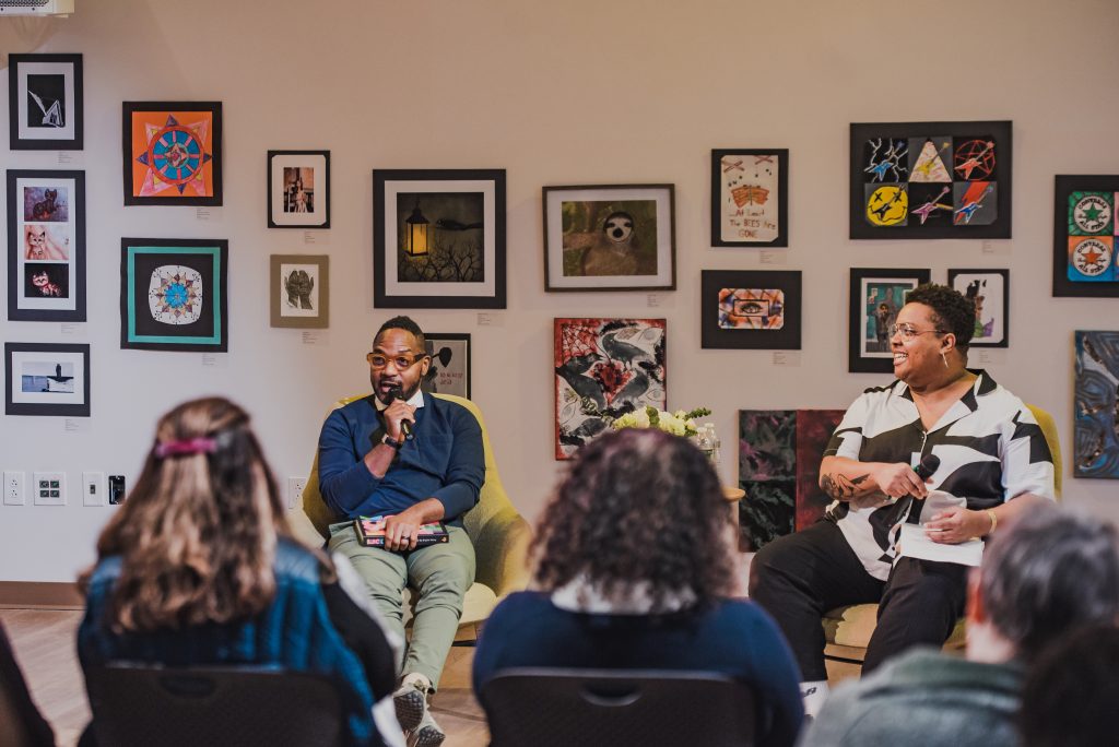 Two speakers sit in a gallery filled with various artworks, addressing an audience during a discussion or event.