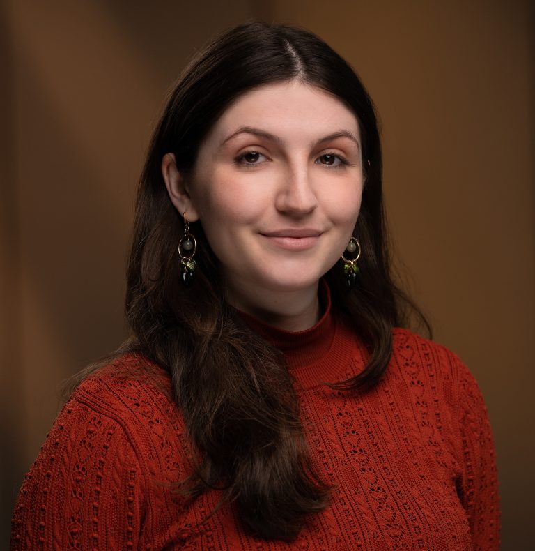 A woman with long dark hair wearing a red sweater and earrings, smiling against a brown background.