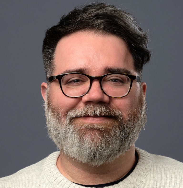 Man with glasses and a beard, smiling against a gray background, wearing a light sweater.
