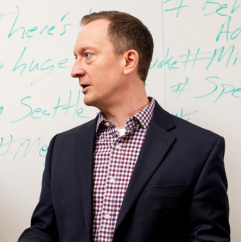 Man with short hair in a blazer, facing sideways in front of a whiteboard with handwritten text.