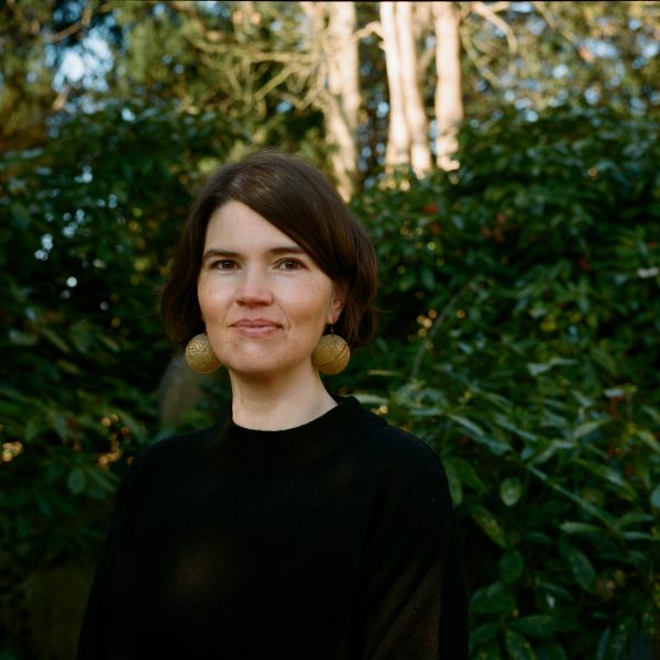 Woman with short brown hair and large earrings, smiling in front of green foliage. Natural light enhances the serene atmosphere.