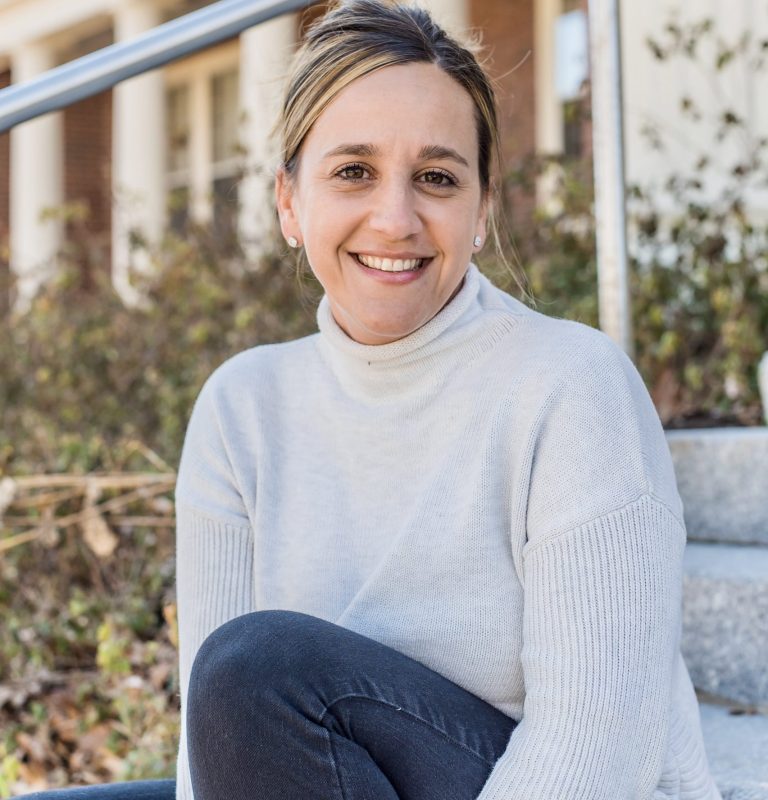 Smiling woman in a light sweater sits on steps outdoors, with greenery and a building in the background.