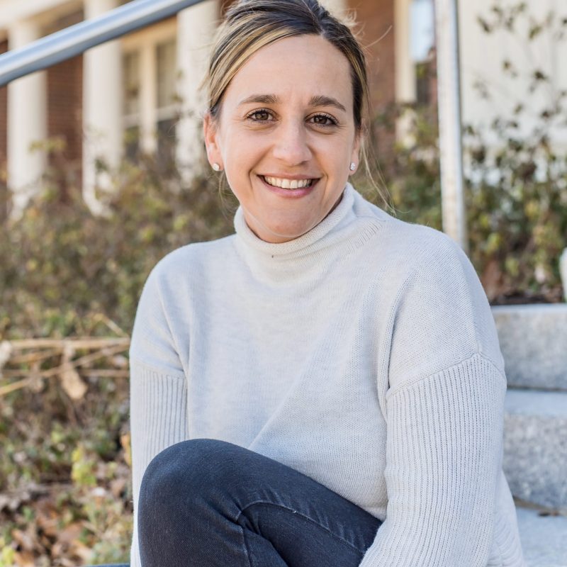 Smiling woman in a light sweater sits on steps outdoors, with greenery and a building in the background.