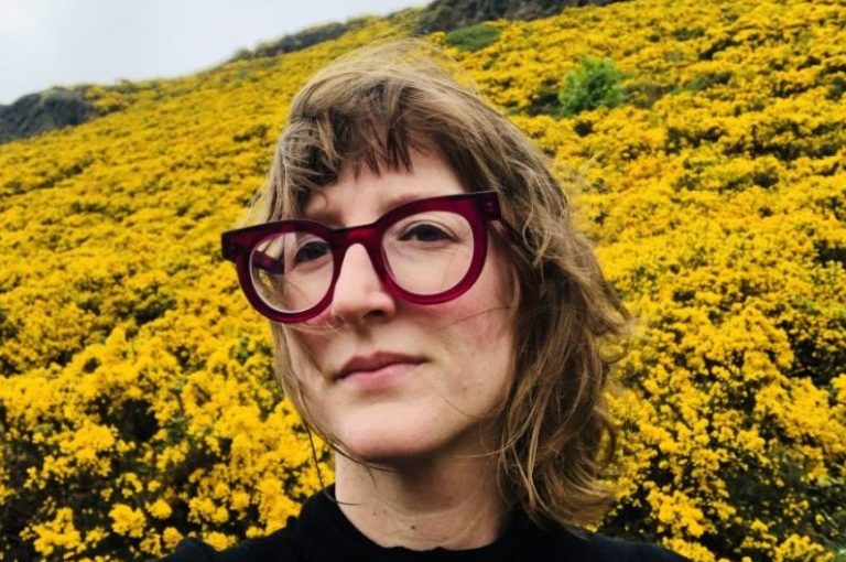 A woman with glasses posing for a photo in front of a field of yellow flowers