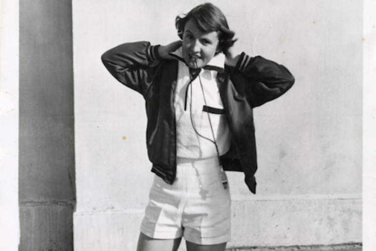 A young woman in a sporty outfit adjusts her jacket, standing against a plain wall. She's wearing shorts and headphones.